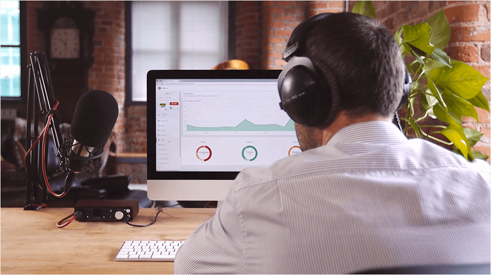 This is a shot of a man from behind wearing a white shirt. He has dark hair and headphones on. There is a Mac screen visible, but what's on the screen is indecipherable. The backdrop is a warm brown office.