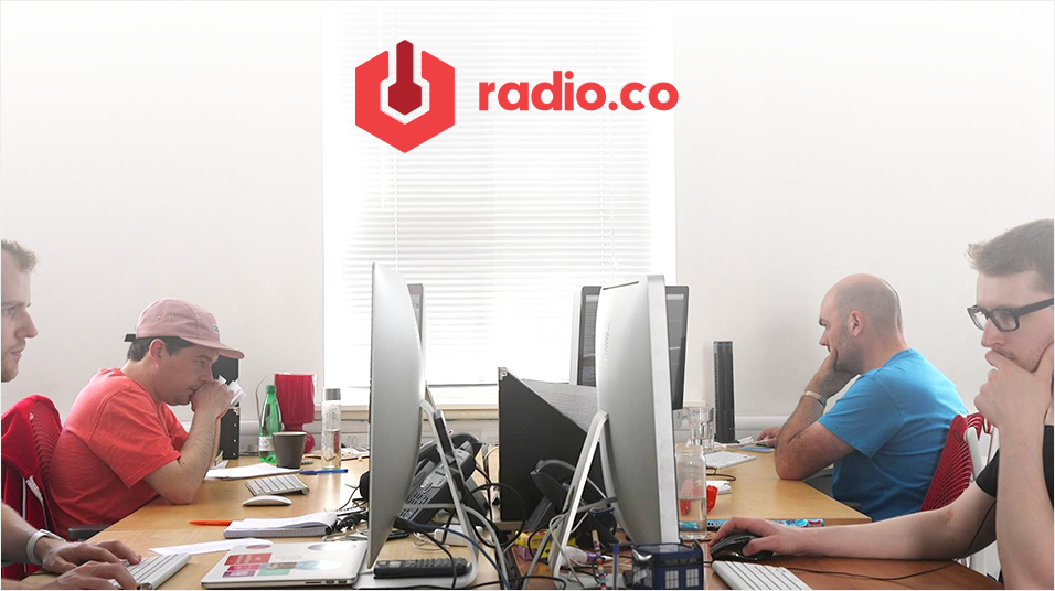 This is a white and red image of the Radio.co offices with the logo at the top. There are computers dividing the middle of the image, and on either side are white mn Mac screens. en with short hair focusing o
