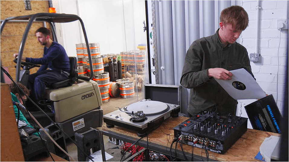 On the right side of the image is a white man with blonde hair putting a record back into its sleeve. he stands in front of a sound mixer and vinyl player. On the left is a white man with dark hair behind the wheel of a moving machine.