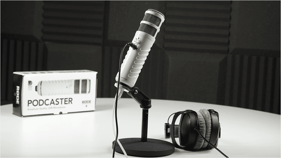The RODE podcaster, on a mic table stand and next to a pair of headphones. The Podcaster box is at the back of the table in the background.
