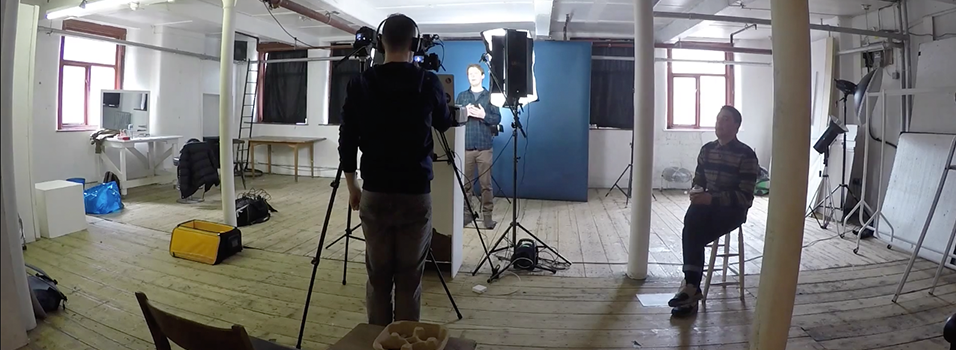 This is an image iof an office with pillars in it. It shows from behind a man wearing a black shirt and short hair listening to headphones. He is filming a man far away talking to the camera in front of a bright light.