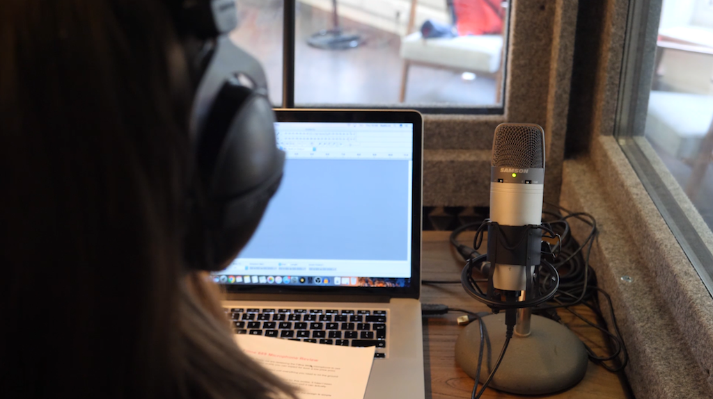 This is an over the shoulder shot of the same person with headphones on looking at a macbook screen. On the right is the microphone in question.