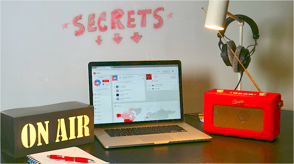 This is an image of a Macbook against a white neutral backdrop on a table. On the right is a Red Roberts radio, above it hangs a mic and a pair of headphones. On the left is an ON AIR lightbox. Above the entire image is a pink graphic of the word 'Secrets