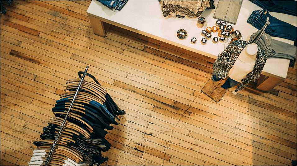 This is an aerial shot of what appears to be a clothes shop. On the bottom left is a rail of dark clothes hung with brown coathangers. The background is a wooden floor. We see the corner of a desk in the top left of the screen.