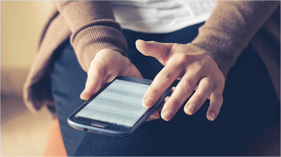 White hands wearing long brown sleeves hold an iPhone. The screen is indecipherable. She uses is over dark denim jeans.