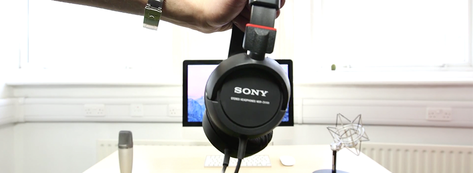 this is the same image as the header image but instead there are black Sony MDR-ZX100 Headphones in front of the monitor here. This is an image with a white backdrop with a silver Mac in the centre. The Mac's screen is a generic purple background. On the left is a silver Mic, on the black is an angular black lamp.