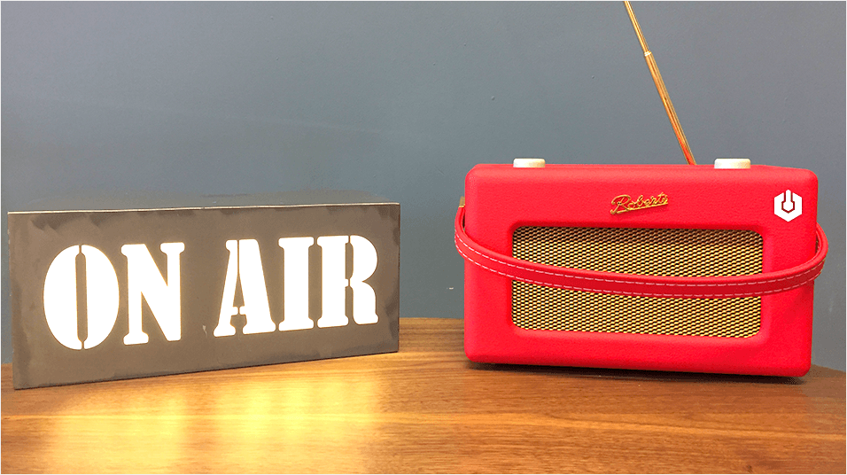 This is an iamge of a red Roberts radio on the right. On the left is a white neon ON AIR sign. The background is grey and neutral.