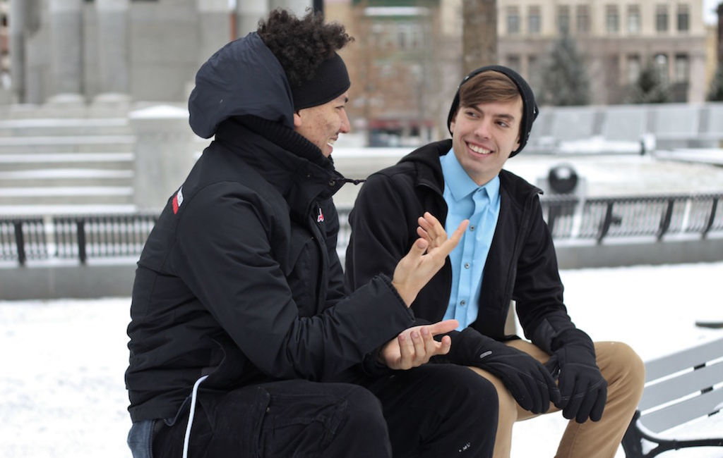 This is a picture of two men in a winter scene talking on a park bench. On the left a man wears a black coat and has black afro hair, and he smiles at his friend on the left who is wearing a blue shirt, black jacket and black beanie over a swept across brown fringe. The two are cheerful.