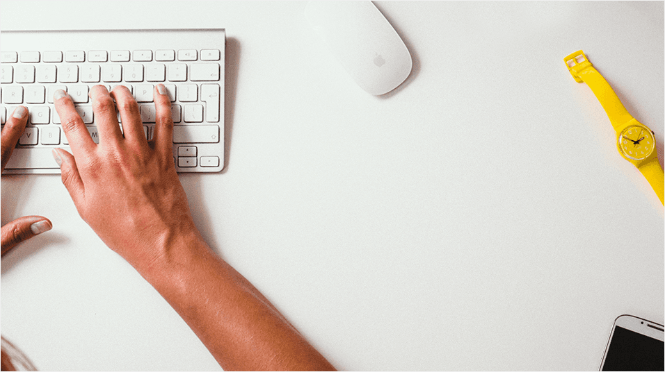 There is a plain white background desk. There is a white arm typing on a Mac keyboard. There is a Mac mouse visible. There is also a yellow wristwatch in shot.