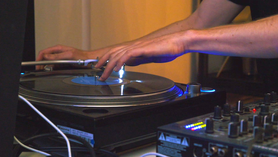 This is an image of a turntable and hands DJing on the turntable. The turntable has blue features and the hands are white.