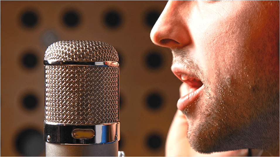 The background is a dotted studio. In the forefront of this picture is a silver mic. On the right hand side is a white man with a shadow beard speaking into the mic.