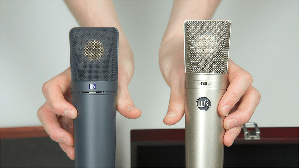 Two white hands holding up the Warm Audio Wa87 against the Neumann U87 to compare them in size. The mic on the right is slightly taller. The background is a plain neutral tone.