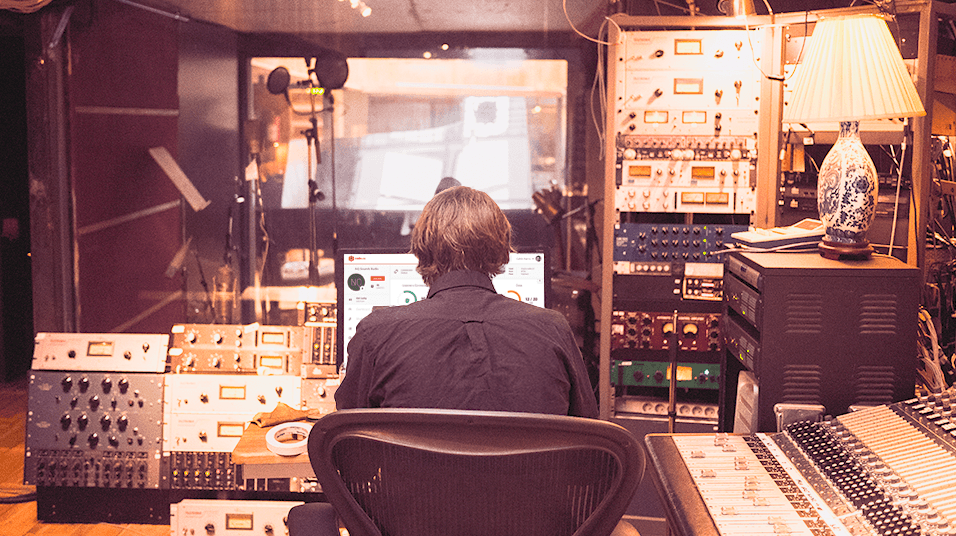 This is an image of a busy radio studio. There is a lamp on the left, warm lighting, and radio mixers and interfaces in silver on the lift. In the centre is a man from behind with greyish hair looking at a computer monitor.